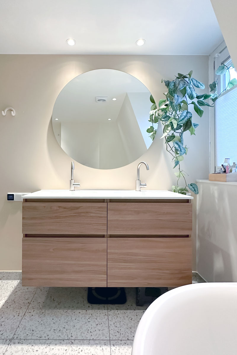 Dubbele wastafel in jaren 30 woning met ronde spiegel en houten ladekast in een vergrote badkamer.